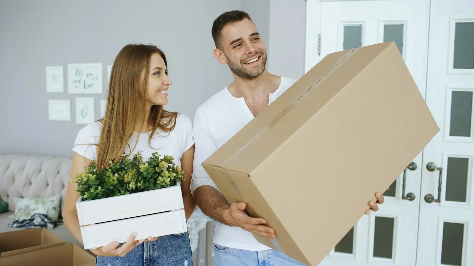 Paar trägt einen Umzugskarton und eine Kiste mit Pflanzen in die neue Wohnung